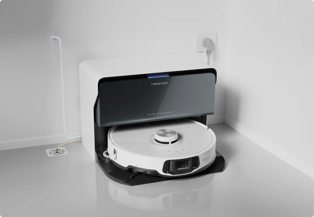 Na obrazie widoczny jest biały robot sprzątający umieszczony w stacji dokującej z napisem \'roborock\'. W tle znajduje się gniazdko elektryczne z podłączonym przewodem.