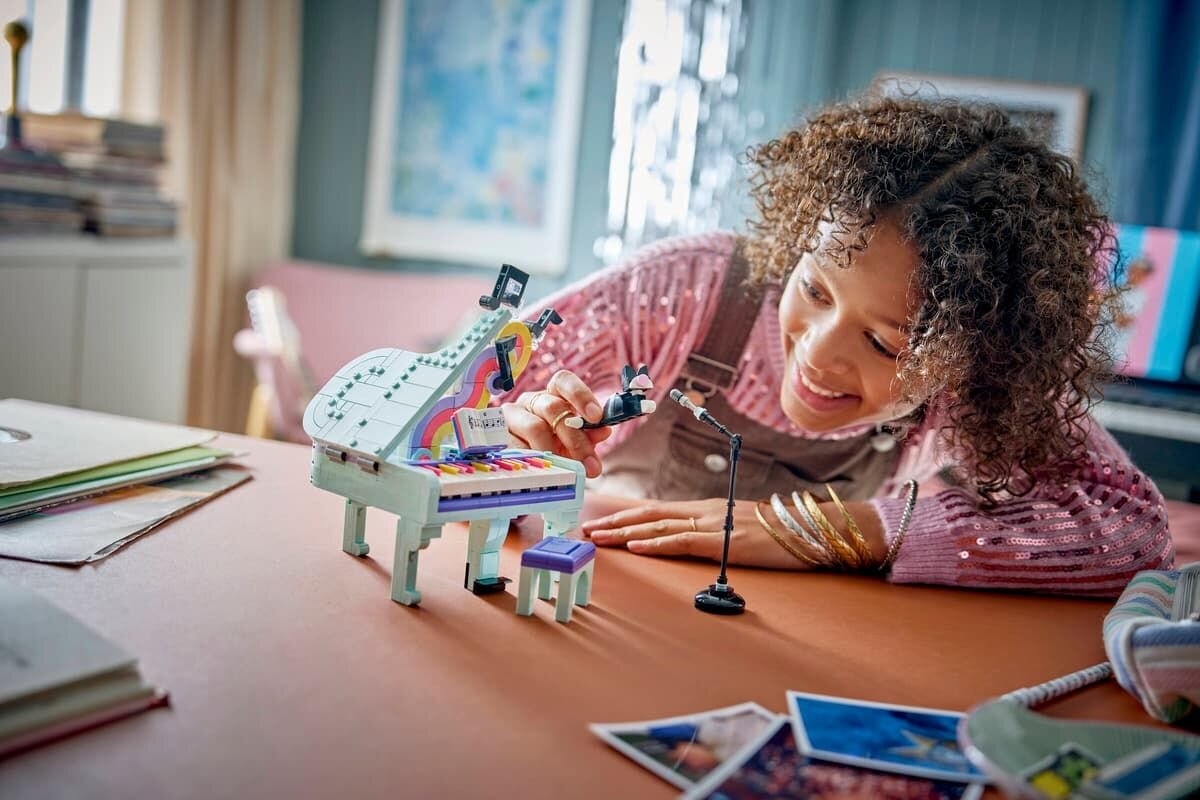 Osoba bawi się kolorowym modelem fortepianu z klocków, na którym znajduje się figurka kota. Obok fortepianu stoi mikrofon na statywie.