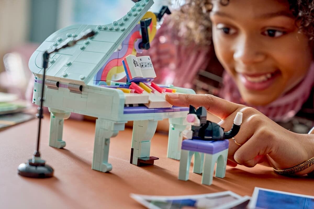 Na obrazie widać model fortepianu z klocków LEGO z kolorowymi klawiszami i nutami na pulpicie. Obok fortepianu stoi figurka kota na stołku, a w tle widoczna jest osoba przyglądająca się zabawce.
