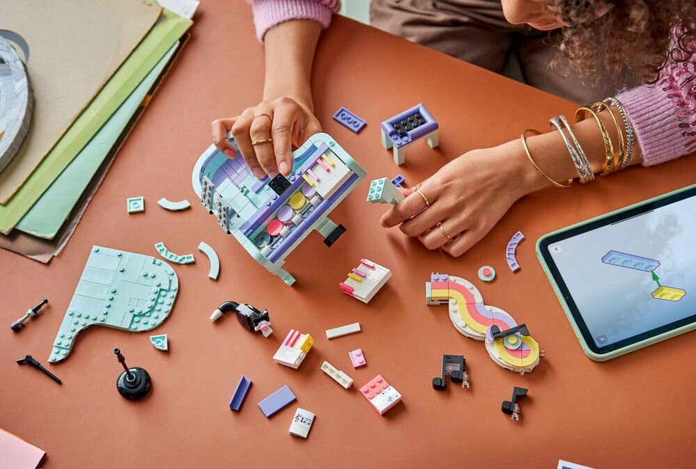 Osoba składa zestaw klocków LEGO na brązowym stole, obok leży tablet z instrukcją. Wokół rozłożone są różnokolorowe elementy LEGO, w tym części przypominające fortepian.