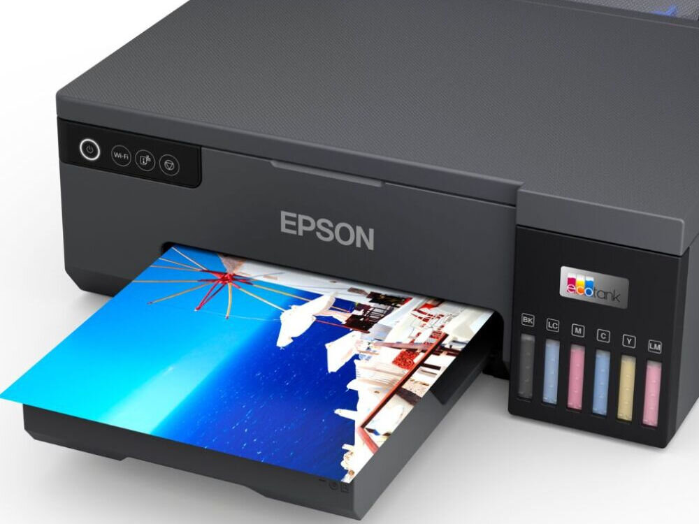 Na obrazie widoczna jest drukarka z logo \'EPSON\', z której wysuwa się kolorowy wydruk przedstawiający krajobraz. Obok znajduje się panel z sześcioma wskaźnikami poziomu tuszu oznaczonymi jako BK, LC, LM, C, Y, M oraz logo \'ecotank\'.