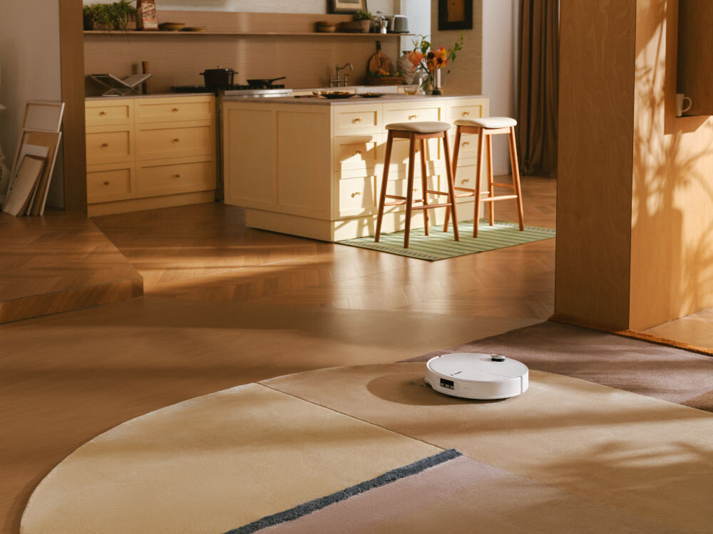 W przestronnym wnętrzu kuchni z drewnianymi szafkami i wyspą kuchenną, na dywanie znajduje się biały robot sprzątający. W tle widoczne są dwa krzesła barowe przy wyspie oraz różne przedmioty na blacie.