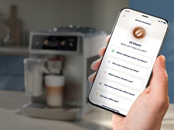 Osoba trzyma smartfon z aplikacją Barista Assistant, na której widoczne są pytania dotyczące kawy. W tle znajduje się ekspres do kawy z filiżanką pod dyszą.