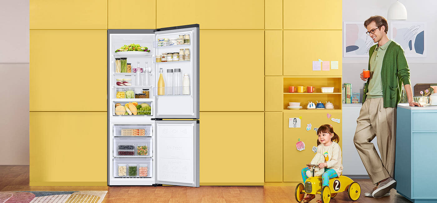 Zdjęcie otwartej lodówki Samsung RB34C671DSA z oferty sklepu Media Expert, która zapewnia elastyczne przechowywanie produktów Na obrazie znajduje się otwarta lodówka wypełniona różnymi produktami spożywczymi, ustawiona na tle żółtej ściany. Obok lodówki stoi mężczyzna z kubkiem oraz dziecko na żółtym rowerku.