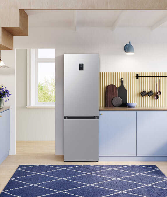Minimalistyczne wnętrze kuchenne, w które wpasowuje się lodówka Samsung RB34C671DSA w kolorze metalicznego grafitu W kuchni znajduje się metaliczna lodówka z wyświetlaczem na drzwiach. Obok niej są niebieskie szafki, a na blacie stoją deski do krojenia i miska.