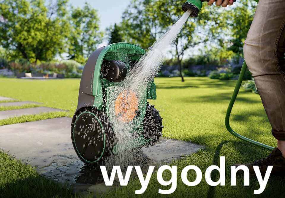 Osoba myje zielony robot koszący wodą z węża ogrodowego na trawniku. Na dole obrazu widoczny jest napis \'wygodny\'.
