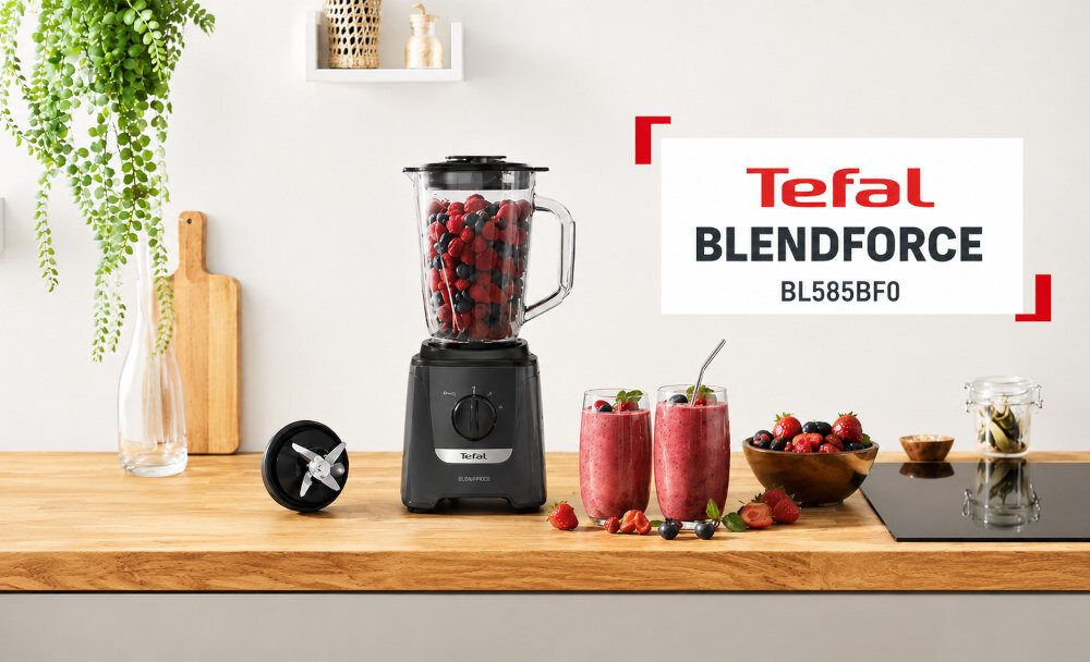 Na drewnianym blacie kuchennym stoi blender kielichowy z logo Tefal, wypełniony czerwonymi owocami. Obok znajdują się dwa szklane kubki z koktajlem oraz miska z owocami, a w tle widoczny jest napis \'Tefal Blendforce BL585BF0\'.