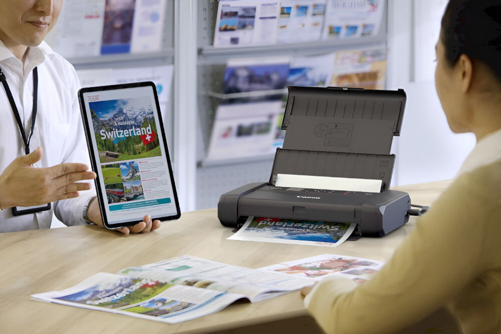 Na stole znajduje się drukarka z otwartą pokrywą, z której wychodzi wydrukowany dokument. Obok osoba trzyma tablet z widocznym tekstem \'Switzerland\'.