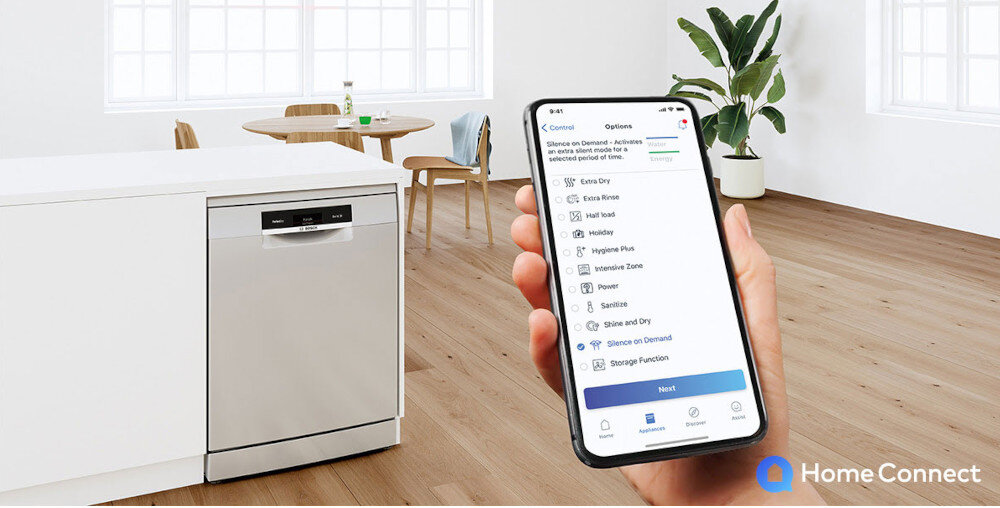 Na obrazie widać zmywarkę w zabudowie kuchennej oraz dłoń trzymającą smartfon z aplikacją Home Connect. W tle znajduje się stół z krzesłami i rośliną w doniczce.