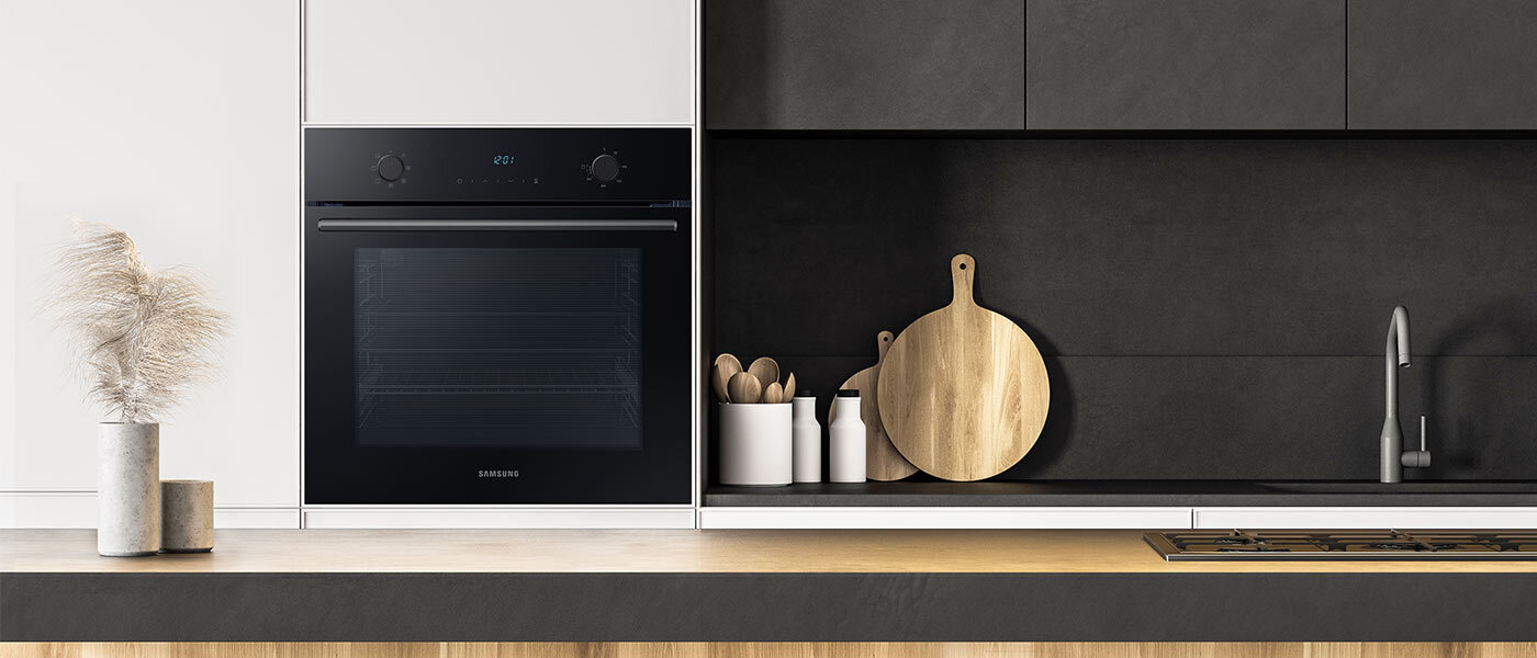 Czarny piekarnik wbudowany w białą szafkę kuchenną, z widocznym logo Samsung. Obok znajduje się ciemna półka z drewnianą deską do krojenia i białymi pojemnikami na przybory kuchenne.