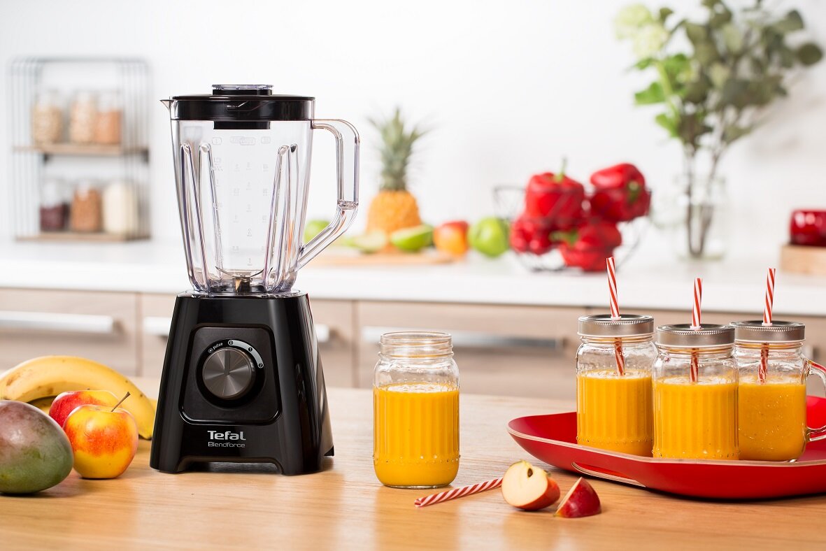 Na blacie kuchennym stoi czarny blender kielichowy z logo \'Tefal BlendForce\'. Obok znajdują się słoiki z pomarańczowym napojem i różne owoce, w tym banan i jabłko.
