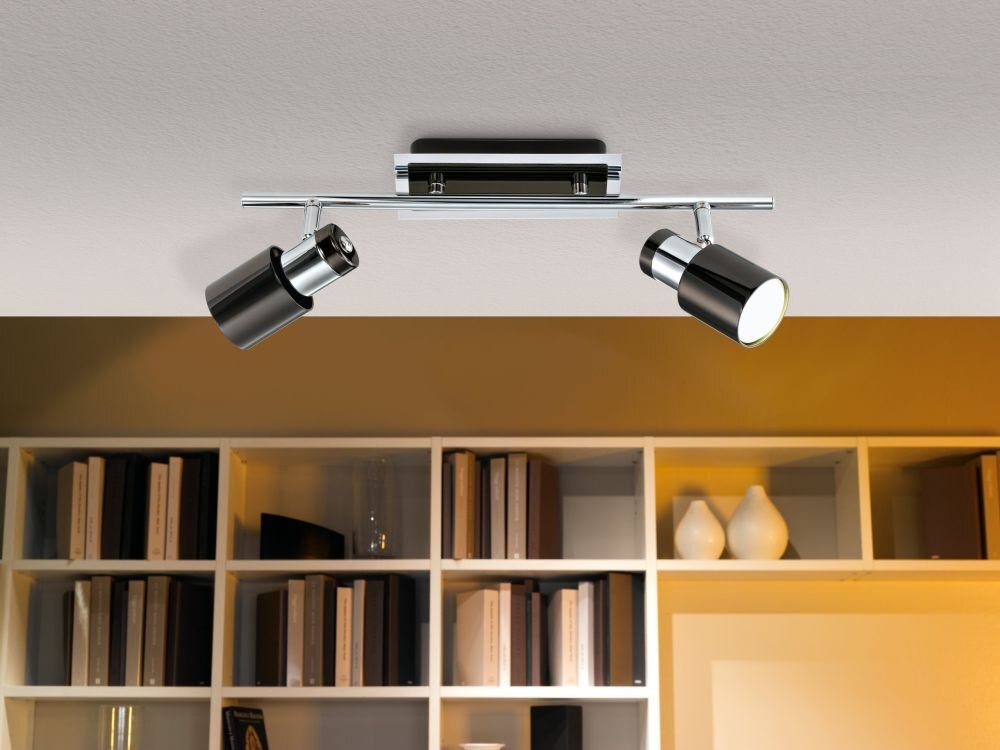 Na suficie zamontowana jest lampa z dwoma czarnymi reflektorami na metalowej listwie. W tle widać półki z książkami i dekoracjami.