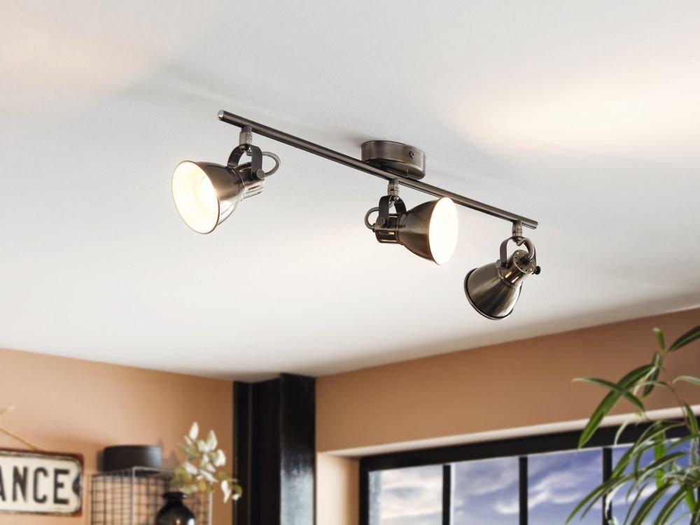 Na suficie zamontowana jest lampa spot reflektorowa z trzema regulowanymi kloszami w kolorze metalicznym. W tle widoczne są elementy wnętrza, takie jak rośliny i fragment okna.