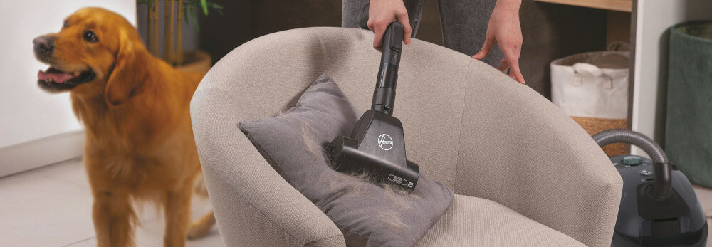 Osoba używa odkurzacza z logo HOOVER do czyszczenia poduszki na fotelu, obok stoi pies. W tle widoczny jest odkurzacz i elementy wnętrza.