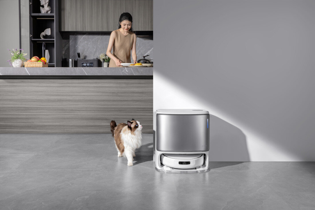 W kuchni znajduje się kobieta przygotowująca posiłek przy blacie, obok stoi robot sprzątający i kot. Robot sprzątający ma nowoczesny, biały design.