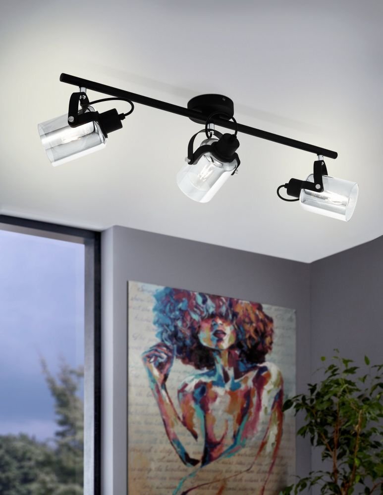 Na suficie zamontowana jest lampa z trzema reflektorami skierowanymi w różnych kierunkach. W tle widoczny jest kolorowy obraz przedstawiający postać oraz fragment okna z widokiem na zewnątrz.