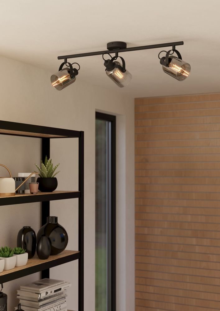 Na suficie zamontowana jest czarna lampa z trzema reflektorami skierowanymi w różnych kierunkach. Obok znajduje się regał z roślinami i dekoracjami oraz okno i ściana z cegieł.