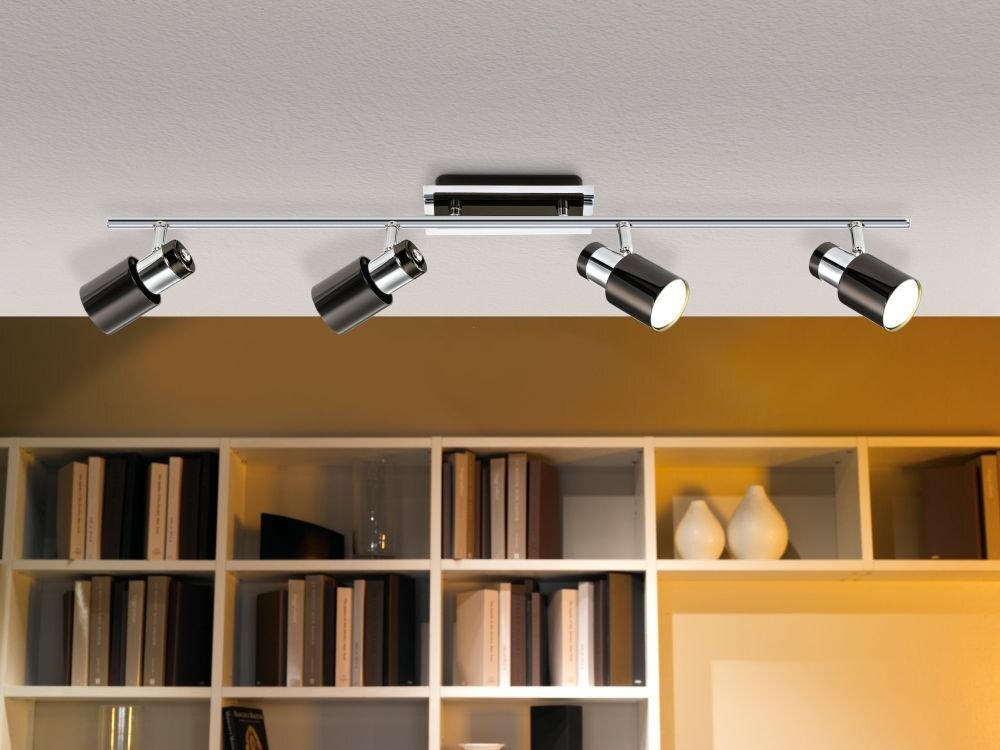 Na suficie zamontowana jest lampa z czterema regulowanymi reflektorami w metalicznym kolorze. W tle widoczny jest regał z książkami i dekoracjami.