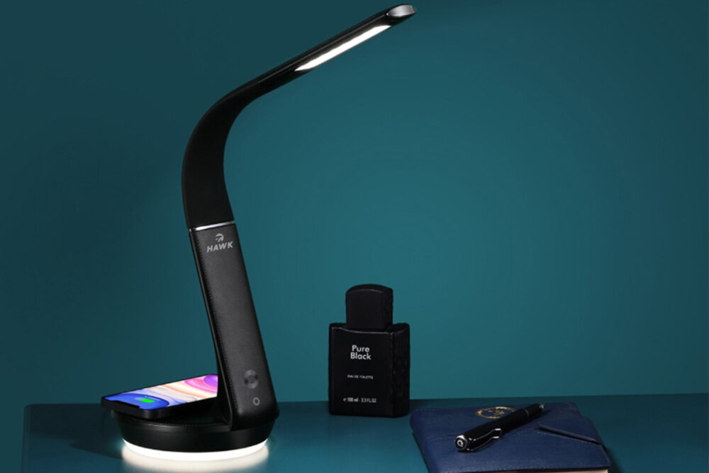 Na obrazie widoczna jest czarna lampka biurkowa z logo \'HAWK\', obok której znajduje się czarny flakon z napisem \'Pure Black\'. Na podstawie lampki leży smartfon, a obok flakonu widoczny jest długopis na notesie.