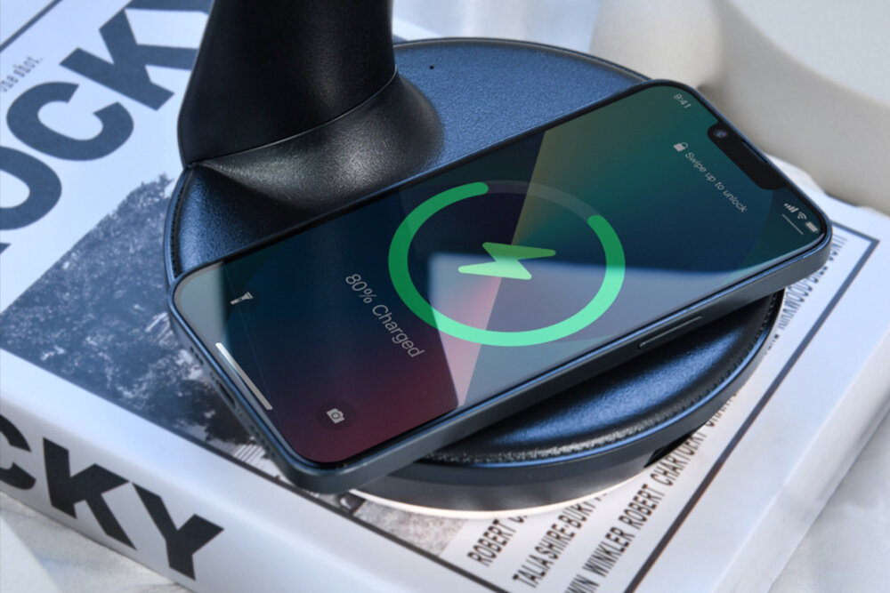 Smartfon leży na bezprzewodowej ładowarce, wyświetlając ekran z informacją \'80% Charged\'. W tle widoczna jest książka z napisem \'ROCKY\'.