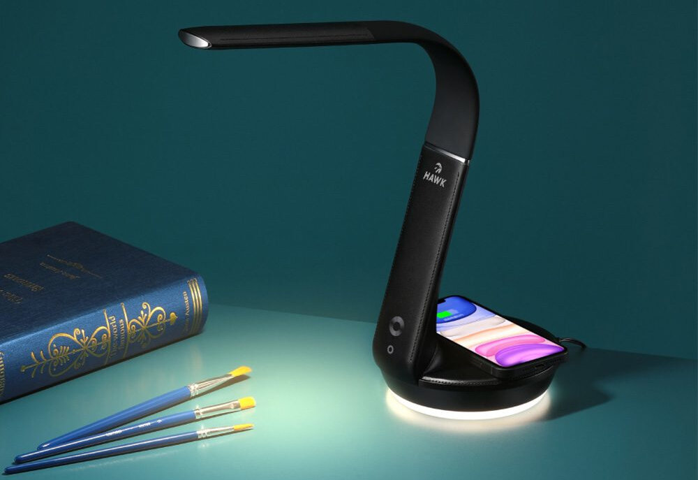 Czarna lampka biurkowa z logo \'HAWK\' oświetla otoczenie, a na jej podstawie znajduje się kolorowy smartfon. Obok leży niebieska książka i trzy pędzle do malowania.