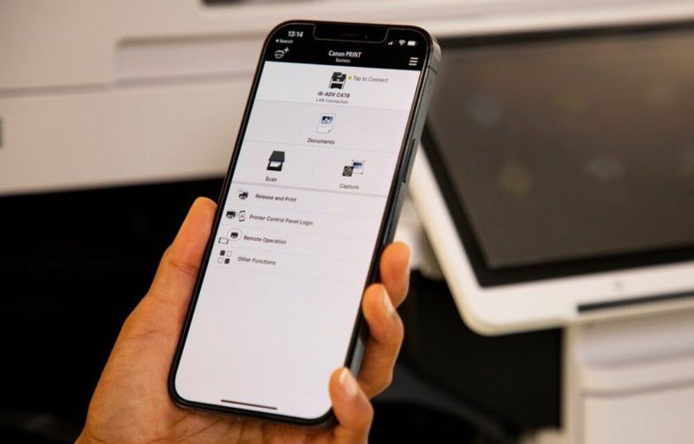 Osoba trzyma smartfon z otwartą aplikacją Canon PRINT, na ekranie widoczne są opcje takie jak \'Scan\', \'Capture\', \'Release and Print\'. W tle znajduje się urządzenie wielofunkcyjne.