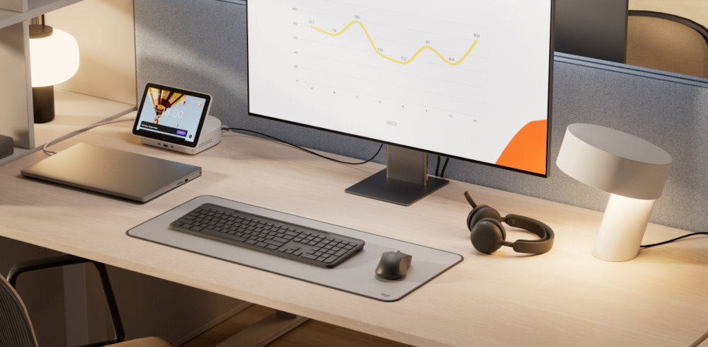 Na biurku znajduje się klawiatura i mysz na podkładce, obok stoi monitor z wykresem oraz lampka i słuchawki. W tle widać laptopa i małe urządzenie z wyświetlaczem.