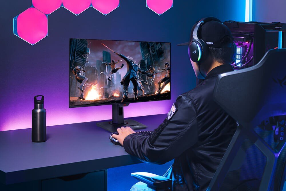 Osoba siedzi przy biurku, grając na komputerze z dużym monitorem, na którym wyświetlana jest dynamiczna scena z gry. Na biurku znajduje się metalowa butelka, a w tle widać podświetlenie LED i komputer stacjonarny.
