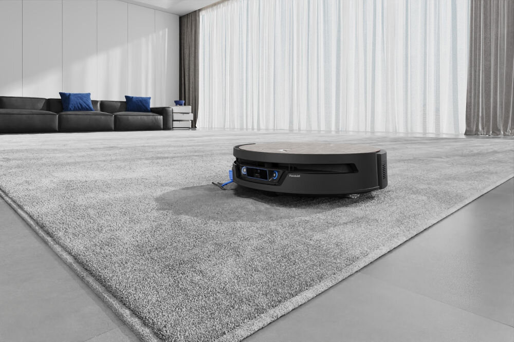 Robot sprzątający znajduje się na szarym dywanie w nowoczesnym salonie z dużymi oknami i zasłonami. W tle widoczna jest czarna sofa z niebieskimi poduszkami.