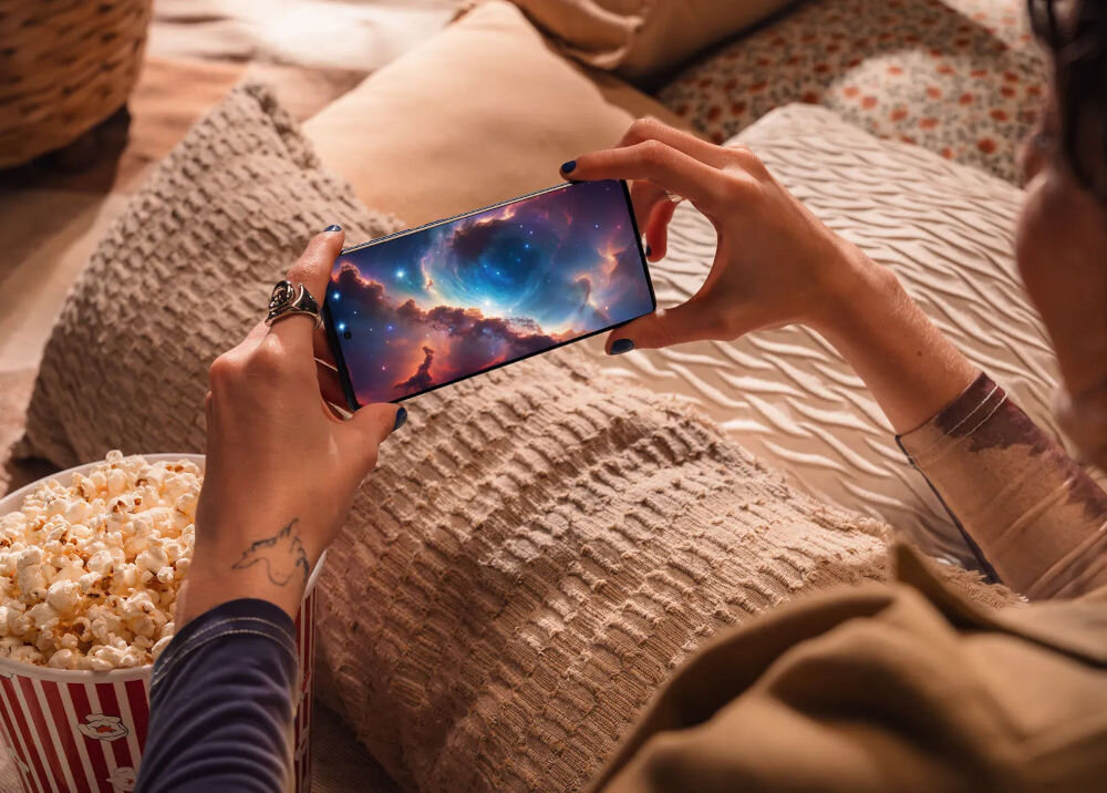Osoba trzyma smartfon, na którym wyświetlany jest obraz kosmosu z kolorowymi chmurami i gwiazdami. W tle widoczne są poduszki i miska z popcornem.
