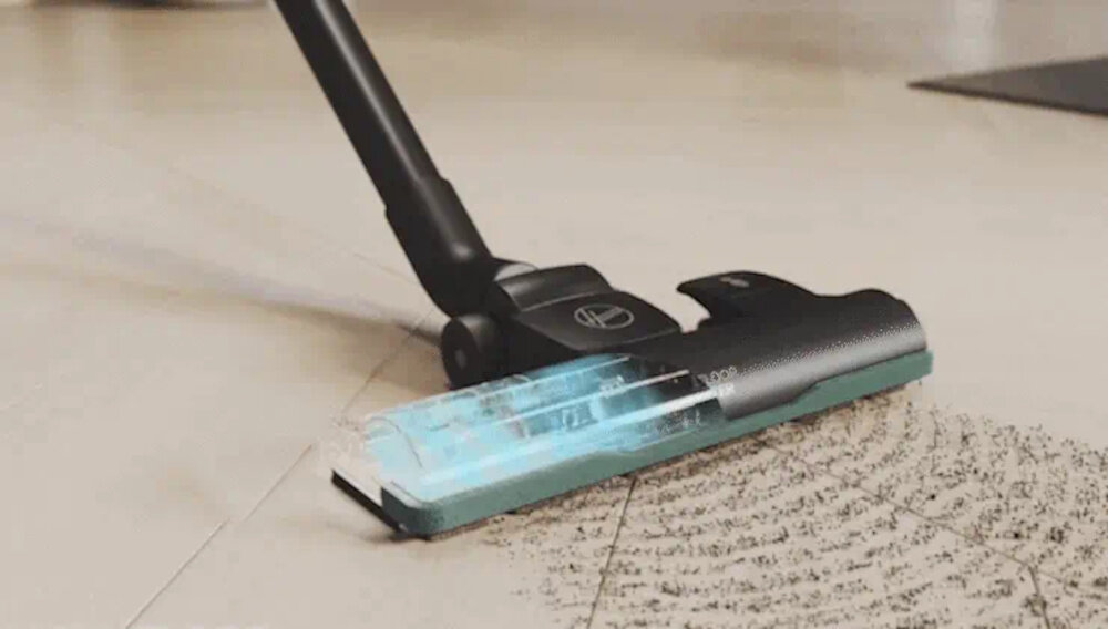 Odkurzacz z logo HOOVER na szczotce, czyszczący podłogę z widocznymi śladami brudu. Szczotka emituje niebieskie światło podczas pracy.