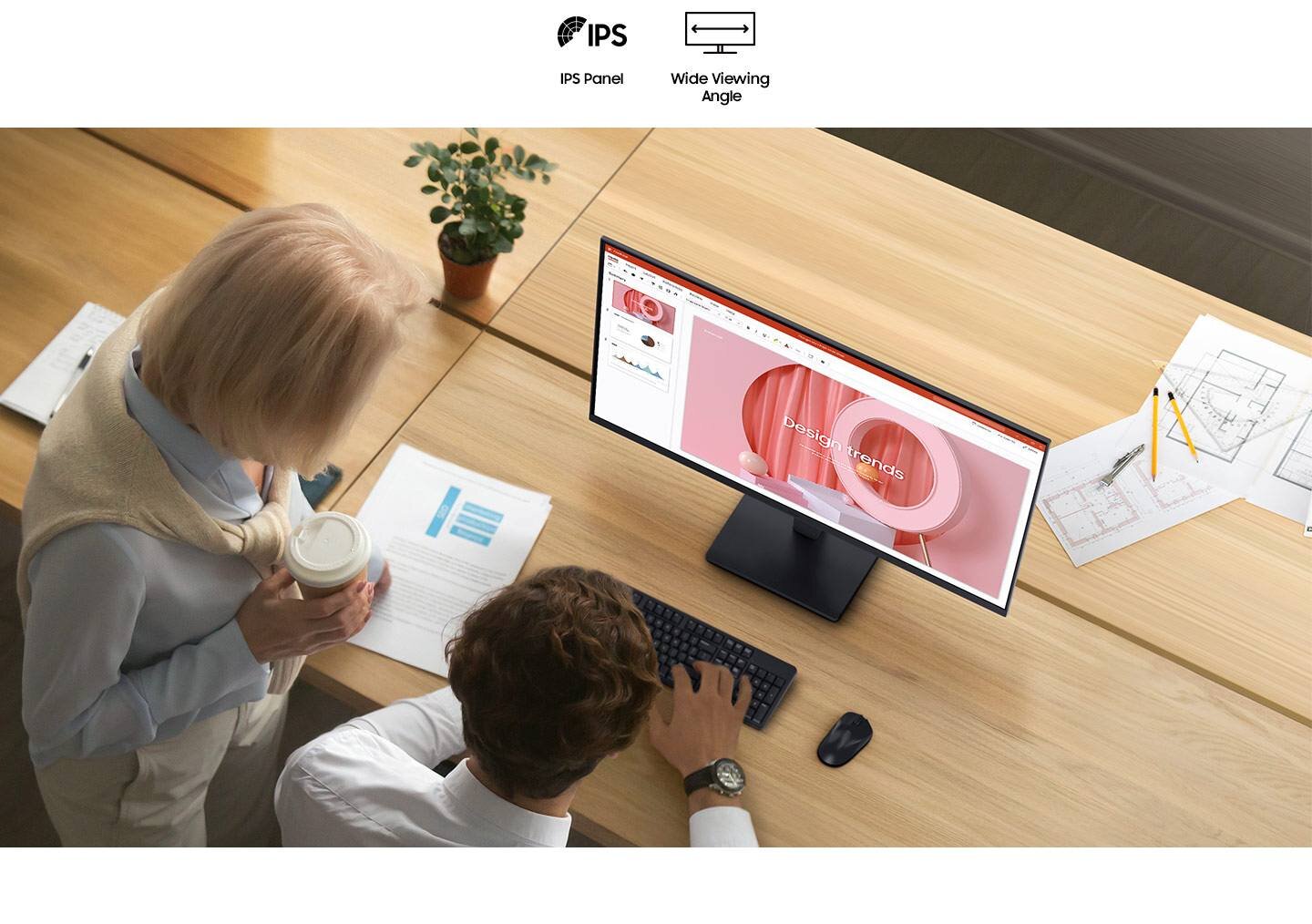 Na biurku znajduje się monitor wyświetlający prezentację z napisem \'Design trends\'. Obok monitora leżą dokumenty i roślina w doniczce, a dwie osoby stoją przy biurku.