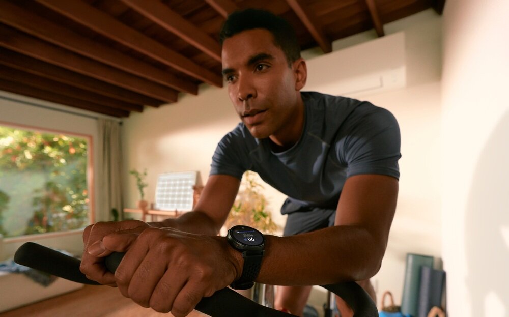 Osoba ćwiczy na rowerze stacjonarnym w pomieszczeniu, nosząc czarny smartwatch na nadgarstku. W tle widać jasne ściany i duże okno z widokiem na zieleń.