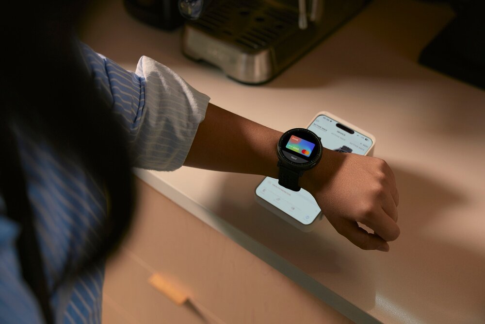 Osoba nosi czarny smartwatch z kolorowym wyświetlaczem na nadgarstku, obok leży smartfon z widocznym ekranem. W tle znajduje się ekspres do kawy na blacie kuchennym.