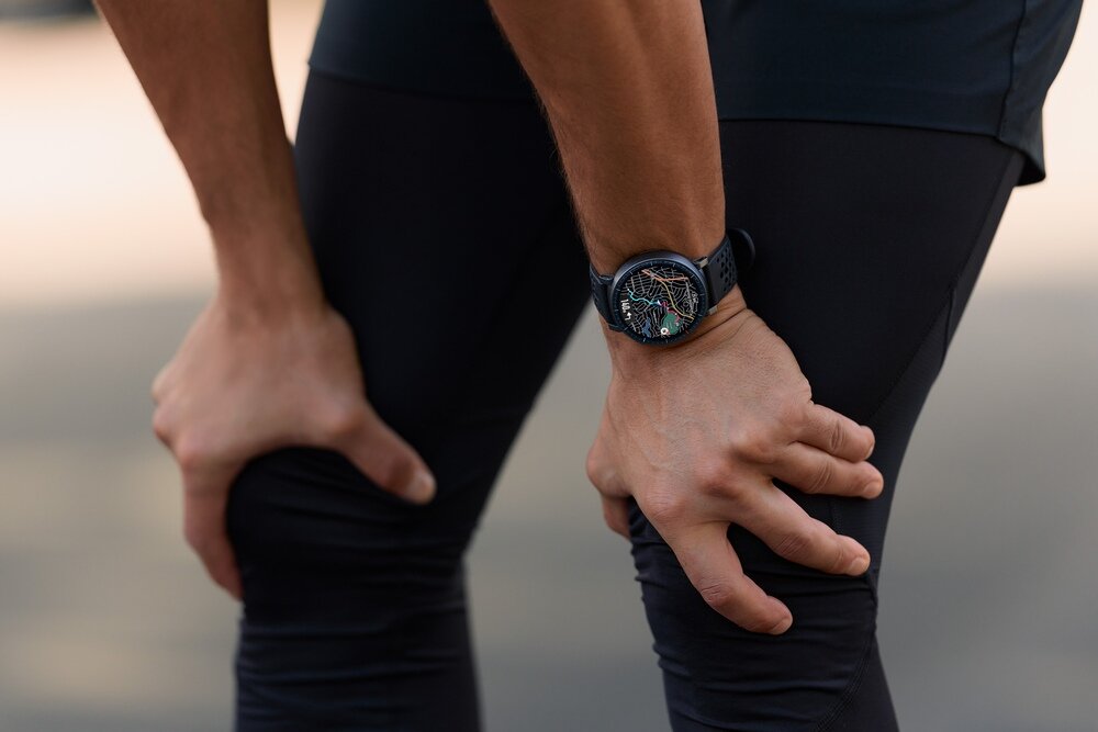 Osoba w ciemnym stroju sportowym trzyma się za kolana. Na nadgarstku widoczny jest czarny smartwatch z kolorowym wyświetlaczem.