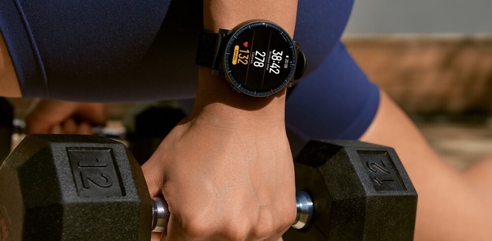 Osoba trzyma hantlę, na nadgarstku ma czarny smartwatch z kolorowym wyświetlaczem pokazującym czas 12:28 i tętno 132. Hantla ma oznaczenie 12 kg.