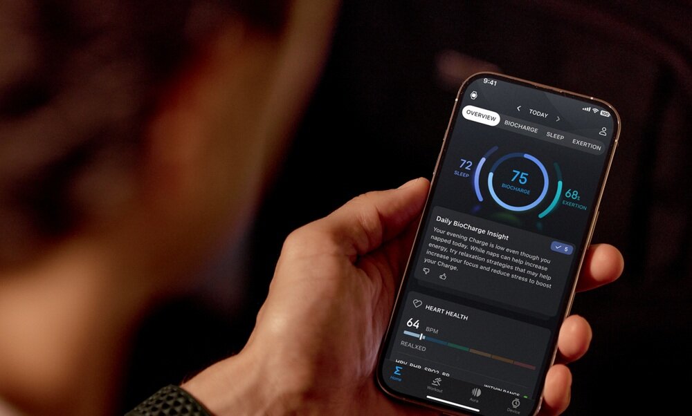 Osoba trzyma smartfon z aplikacją wyświetlającą dane zdrowotne, takie jak \'BioCharge\' z wartością 75. Na ekranie widoczne są także sekcje dotyczące zdrowia serca i snu.