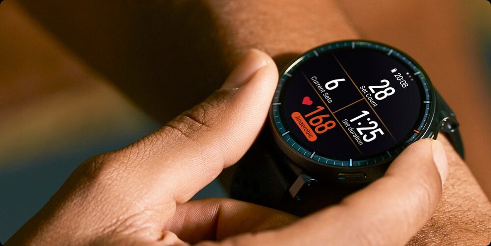 Osoba nosi czarny smartwatch z okrągłym wyświetlaczem, na którym widoczne są dane dotyczące kroków, tętna i czasu. Na ekranie widnieje tekst: \'20:08\', \'6\', \'28\', \'168\', \'1:25\'.
