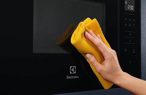 Osoba czyści czarną powierzchnię kuchenki mikrofalowej żółtą ściereczką. Na urządzeniu widoczne jest logo Electrolux oraz wyświetlacz z godziną 2:30.