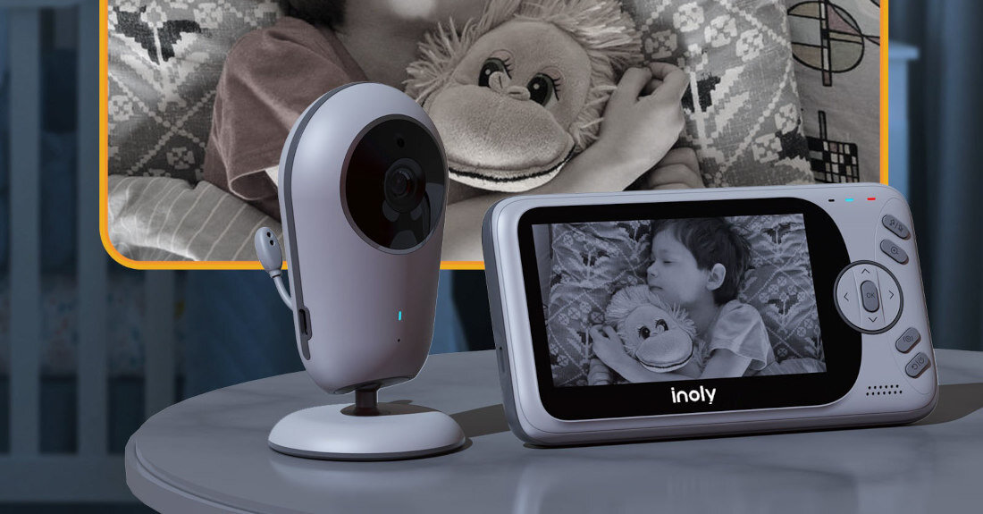 Na stole znajduje się elektroniczna niania z kamerą i monitorem, na którym widać dziecko śpiące z pluszakiem. Na monitorze widnieje logo \'inoly\'.