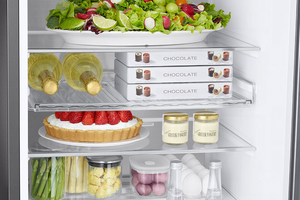 schłodzisz butelki z napojami i produkty w plastikowych lub kartonowych opakowaniach Wnętrze lodówki z widocznymi półkami, na których znajdują się różne produkty spożywcze, w tym sałatka, butelki wina, pudełka z napisem \'CHOCOLATE\', ciasto z truskawkami, jogurty oraz warzywa w słoikach i pojemnikach.