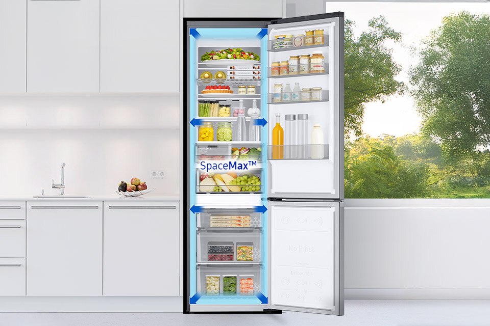 Lodówka RB38C7AGWS9/EF z technologią SpaceMax Otwarta lodówka z widocznymi półkami wypełnionymi różnorodnymi produktami spożywczymi, na drzwiach napis \'SpaceMax™\'. W tle znajduje się jasna kuchnia i widok na zieleń za oknem.
