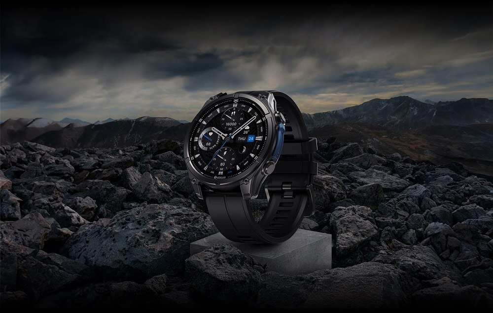 Czarny smartwatch z okrągłą tarczą umieszczony na kamienistym tle z górskim krajobrazem w tle. Na tarczy widoczne są wskazówki, cyfry oraz dodatkowe wskaźniki.