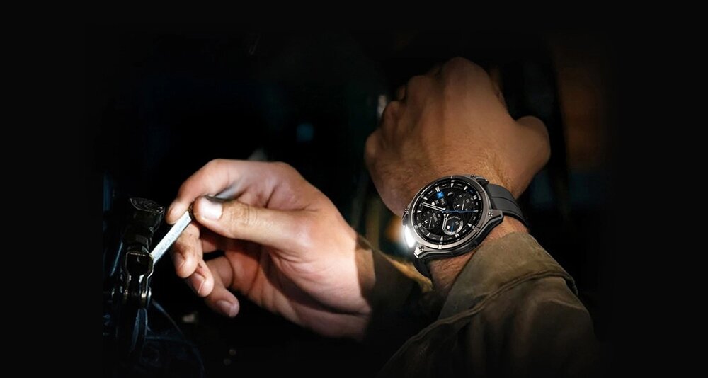 Osoba nosi czarny smartwatch z okrągłą tarczą i widocznymi wskazówkami. Obok trzyma klucz w dłoni, w ciemnym otoczeniu.