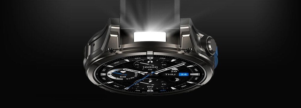 Obraz przedstawia czarny smartwatch z okrągłym wyświetlaczem, na którym widoczny jest analogowy zegar z dodatkowymi wskaźnikami. Tło jest ciemne, a nad zegarkiem znajduje się jasne światło.