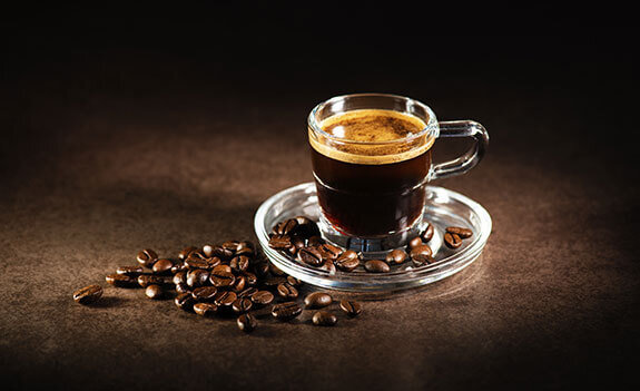 Szklanka espresso na szklanym spodku, obok rozsypane ziarna kawy na ciemnym tle.