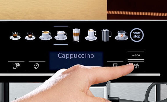 Panel dotykowy ekspresu do kawy z ikonami różnych napojów, w tym cappuccino, oraz przyciskiem \'start stop\'. Widoczna jest dłoń wskazująca na ekran.