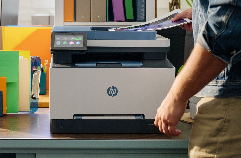 Na biurku znajduje się urządzenie wielofunkcyjne z logo HP, obok którego leży kilka kolorowych segregatorów. Osoba trzyma wydrukowany dokument nad urządzeniem.