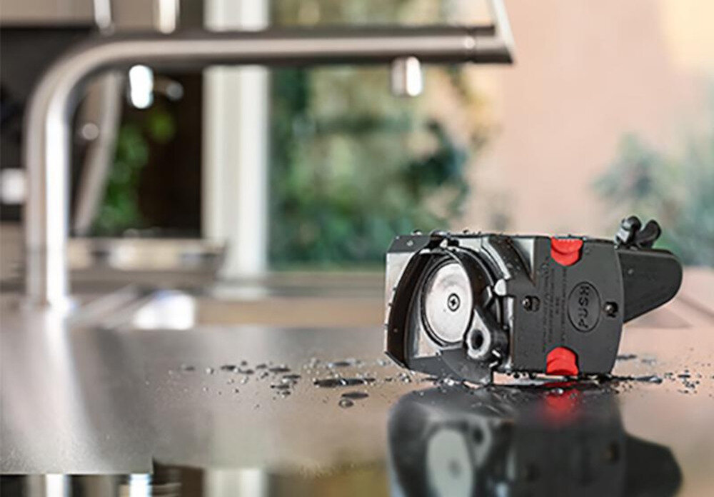 Na blacie kuchennym leży część ekspresu do kawy z widocznymi elementami mechanicznymi i czerwonymi przyciskami. W tle znajduje się kran i rozmyte tło kuchenne.