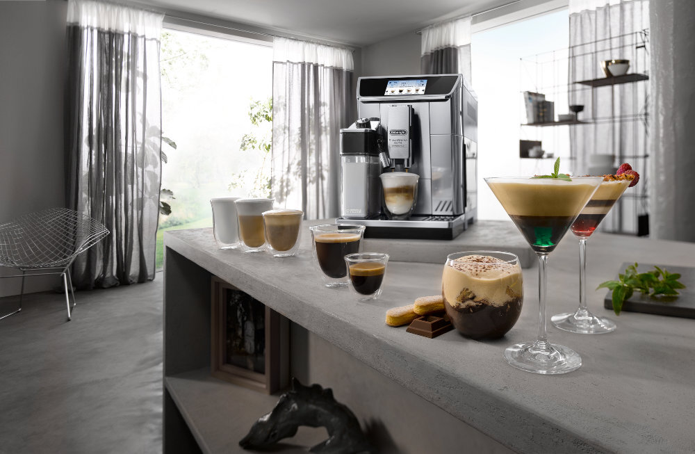Na blacie kuchennym stoi ekspres do kawy z logo \'DeLonghi\', obok którego znajduje się kilka filiżanek i szklanek z różnymi napojami kawowymi. W tle widać jasne zasłony i nowoczesne wnętrze.